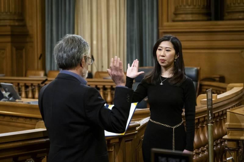 An Asian woman wearing a black dress raises her right hand in front of a man wearing a black suit who is also raising his and holding a document inside a building.