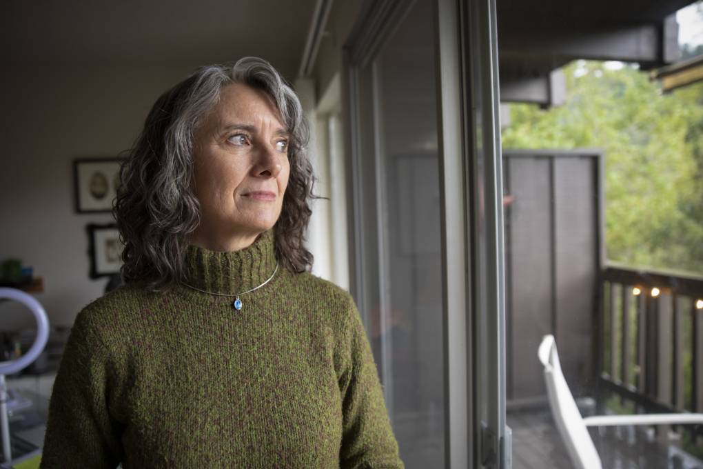A white woman wearing a dark green sweater and necklace looks out a window.