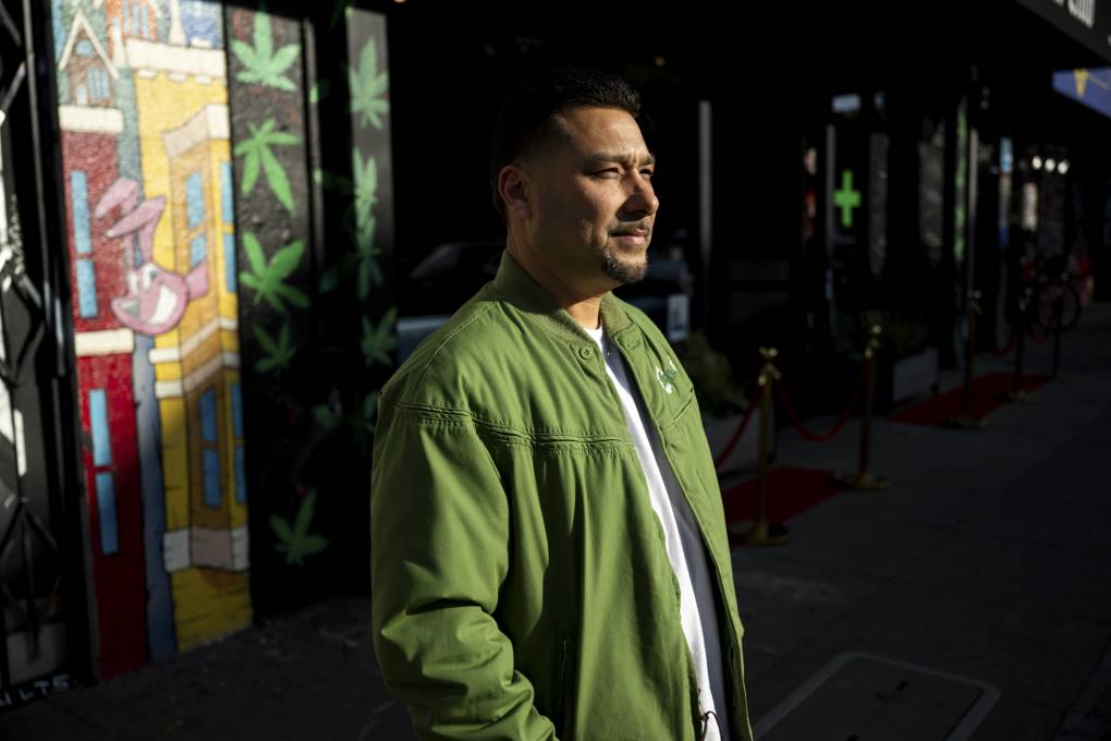 A man wearing a green bomber jacket stands near a wall with cannabis leaves in the background.