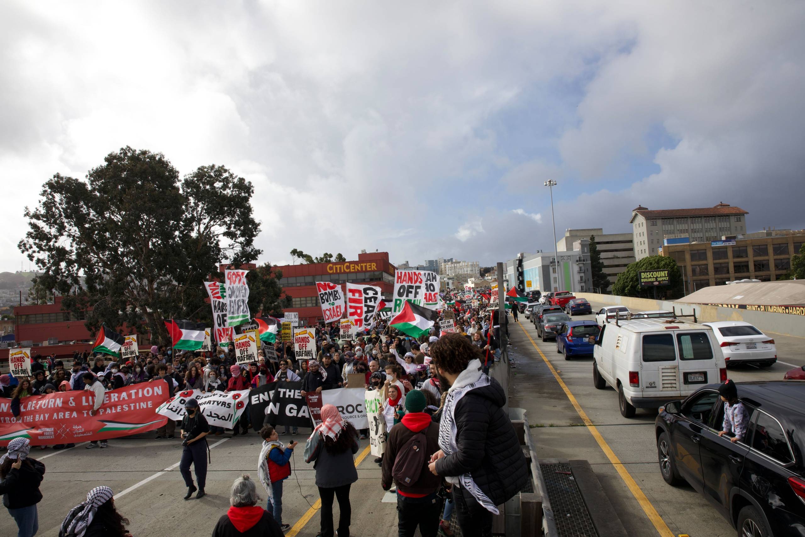 A crowd carrying signs blocks half a roadway, with cars backed up on the other side of the divider.