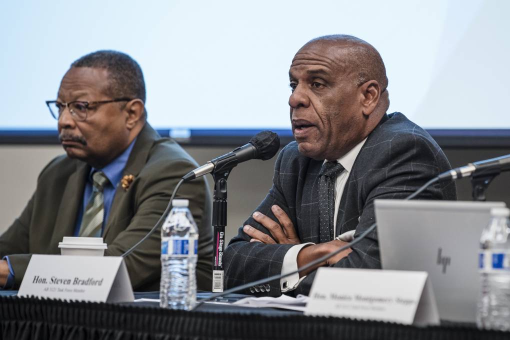 Two Black men in suits sit at a table with microphones, speaking to a room.