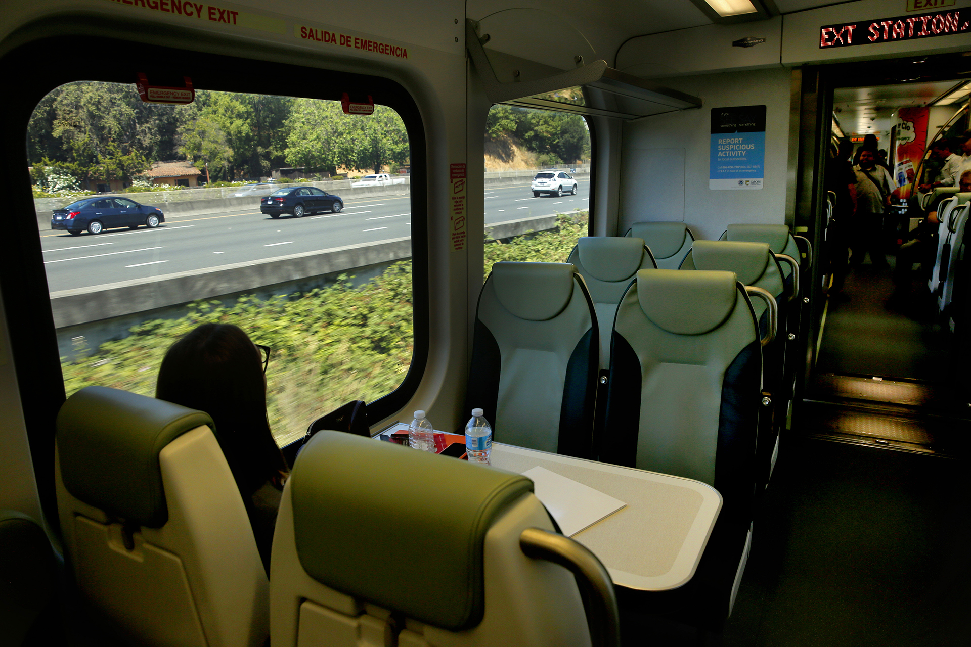 A woman sitting on a train looks out a window and Highway 101, which is running parallel to the train she is on. There are a few cars on the roadway.