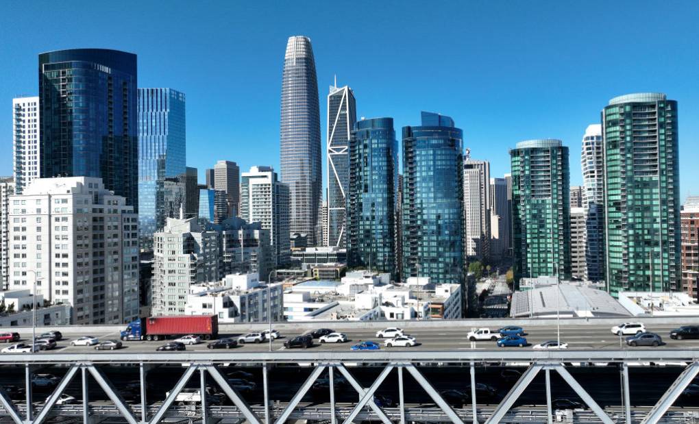 A city skyline with high rises and a blue sky, cars driving in foreground on a bridge.