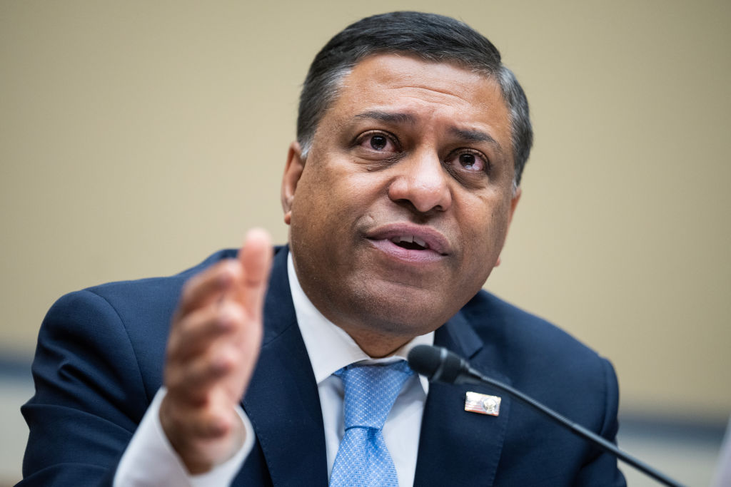 A seated Indian man wearing a suit and blue tie speaks into a microphone.