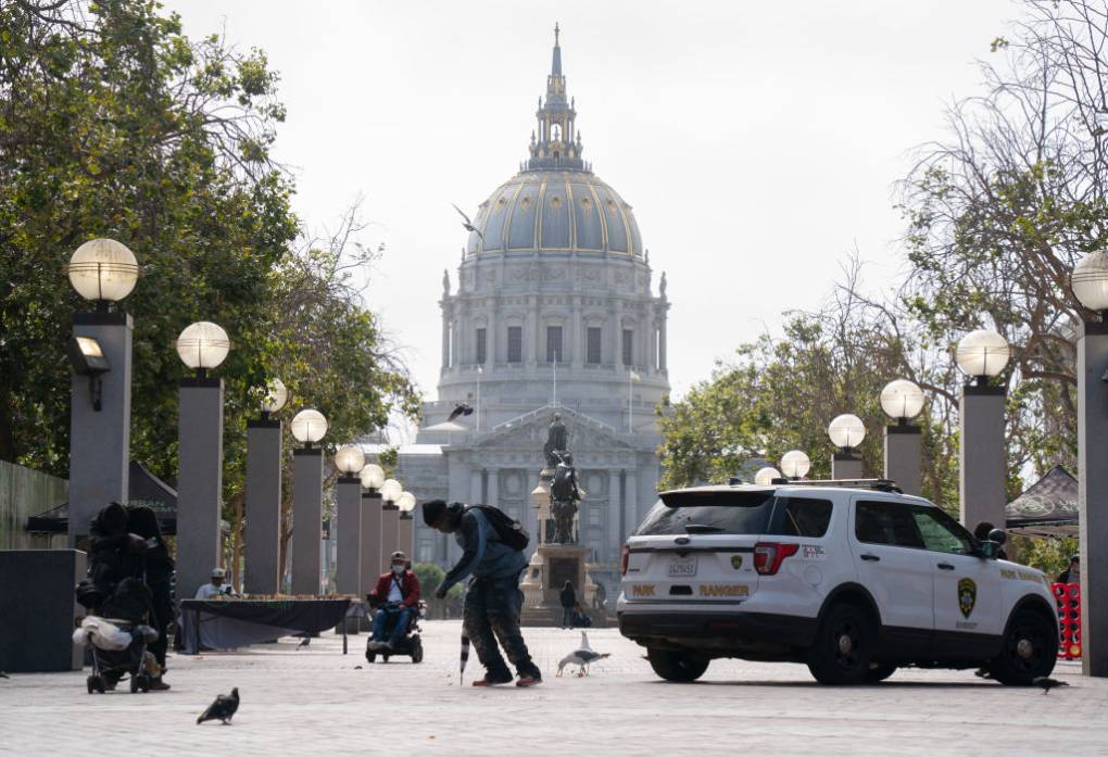 A stopped-over man walks past San Francisco City Hall, with a law enforcement vehicle parked nearby in the plaza.