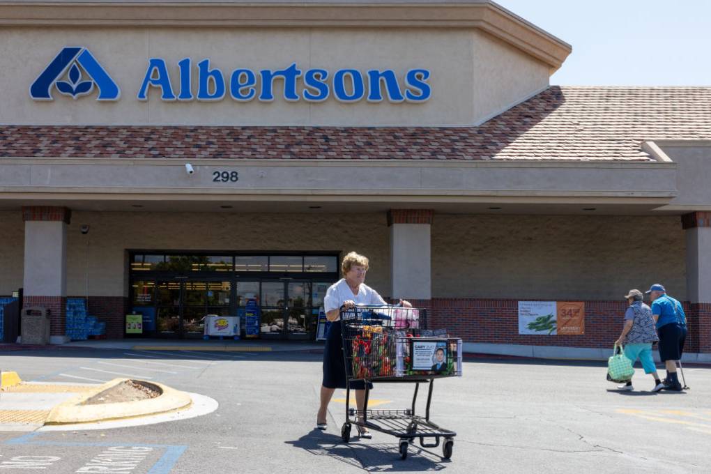 An elderly woman pushes a shopping cart in a parking lot in front of an Albertsons supermarket.