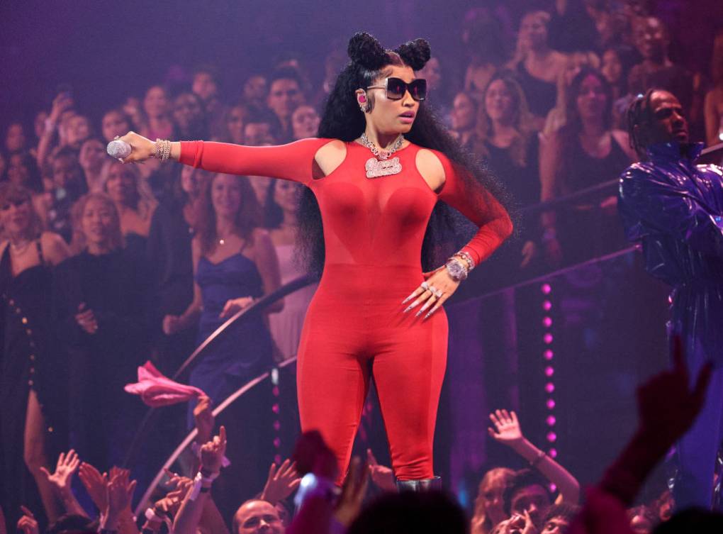 A female singer in a red body suit and sunglasses extends the mic to the crowd while glancing to the side, surrounded by an audience in mid-performance.