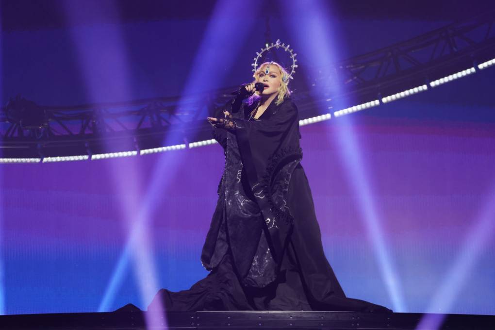 A white woman dressed in an elaborate black gown and wearing a halo crown holds a microphone on stage.