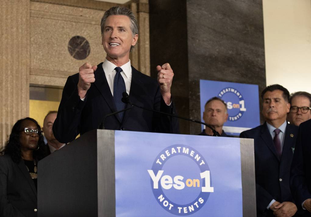 A silvery-haired man with a black suit stands at a podium with a "Yes on 1" banner