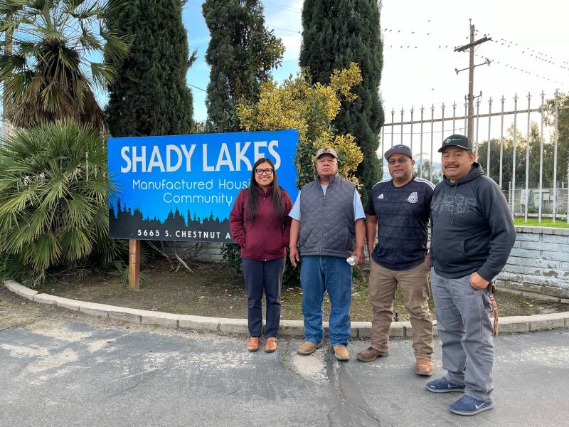 Three men and one woman stand in front of a sign that reads "Shady Lakes" next to some trees outside.