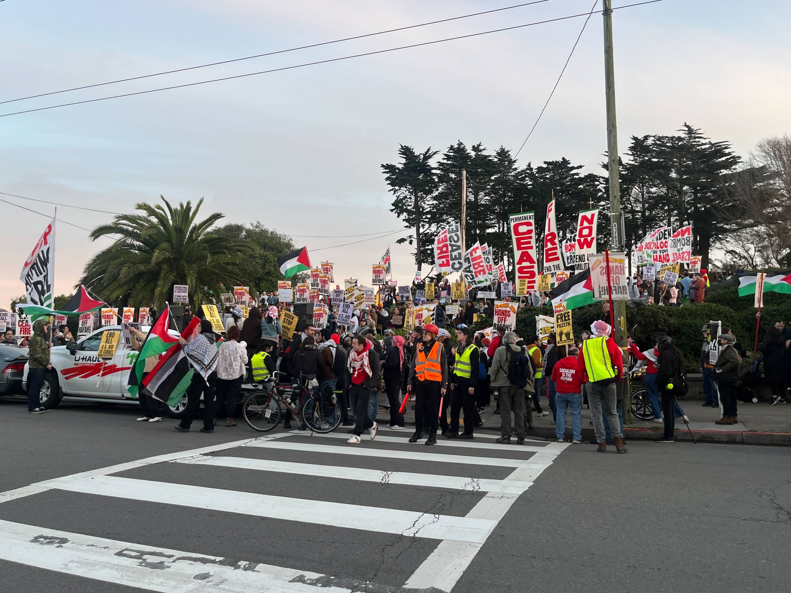 Protesters by the side of a road with a park behind them holding banners and chanting.
