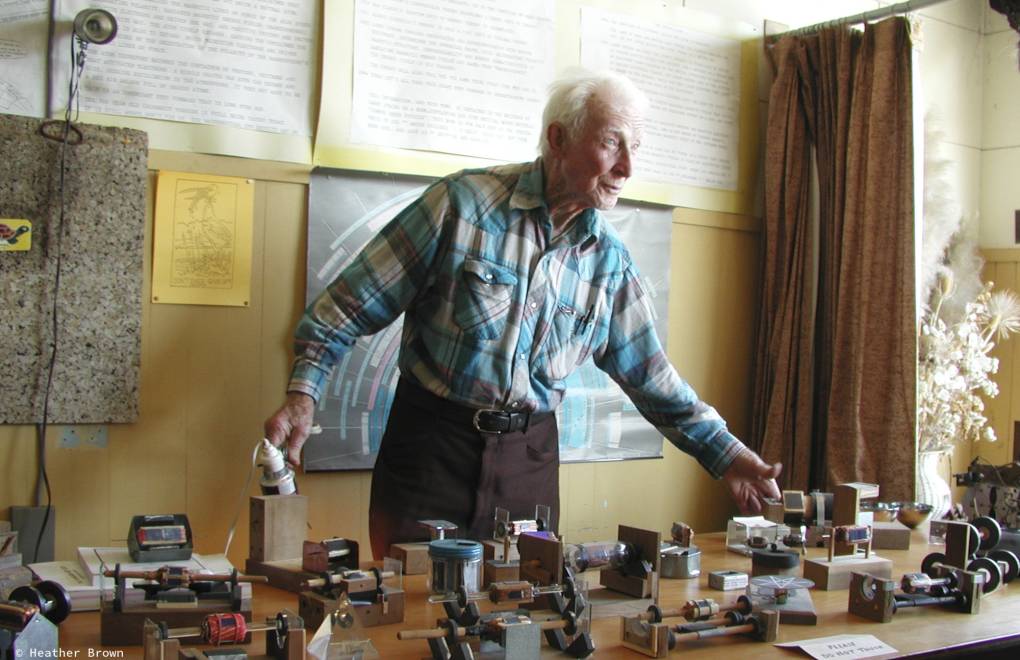 An older man wearing a plaid top stands in front of a yellow wall with a table of motors.