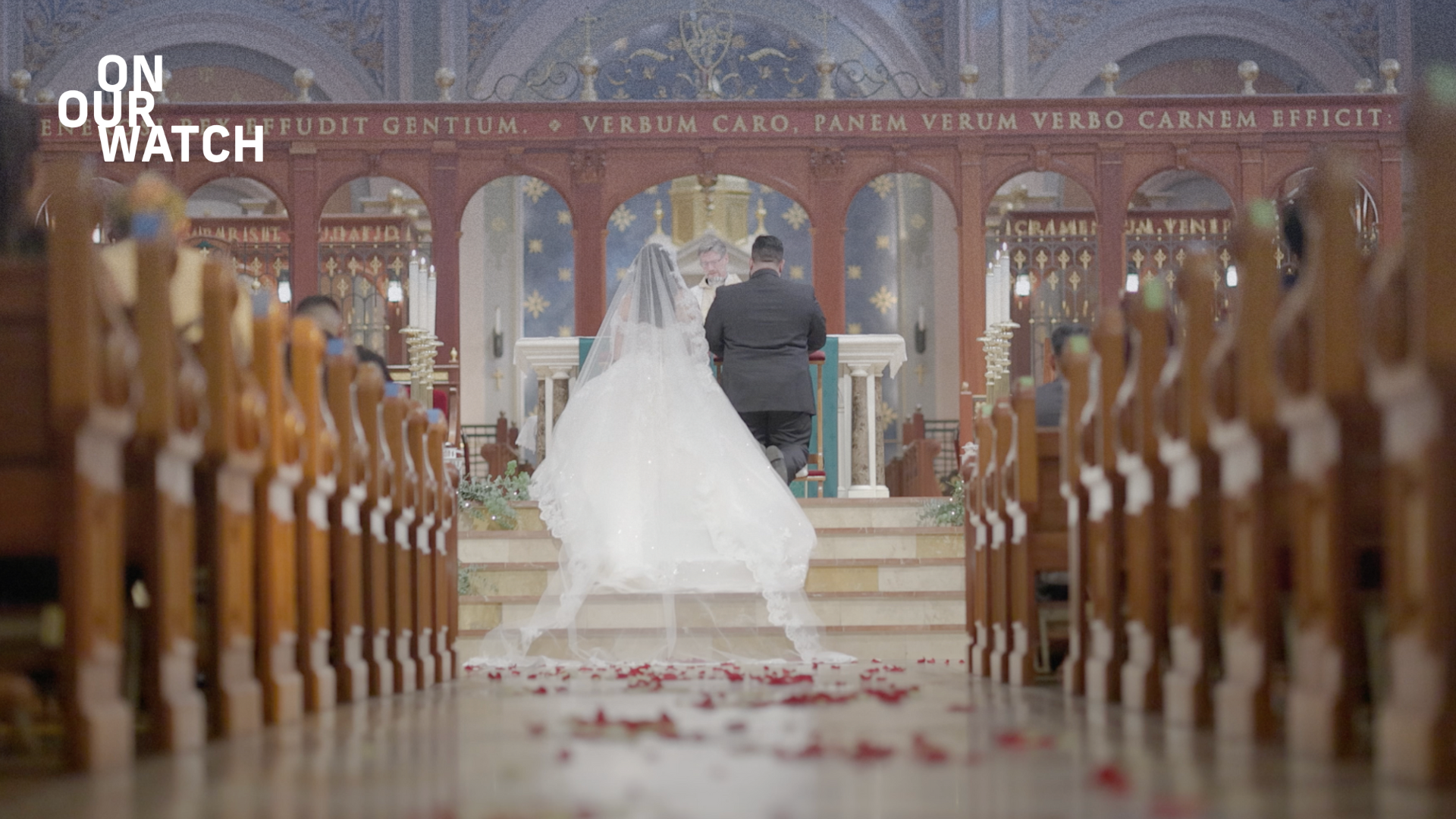 A man wearing a dark suit and a woman wearing a white wedding dress stand next to each other on the steps of a dais inside a cathedral. They are facing with their backs towards the camera, the man on the left and the woman on the right. The cathedral is ornately decorated with gold crosses, latin words embossed in gold stretching across a wooden beam and archway above the couple. An officiant stands before them, a man with gray hair and goatee and glasses. The wooden pews are filled with people.