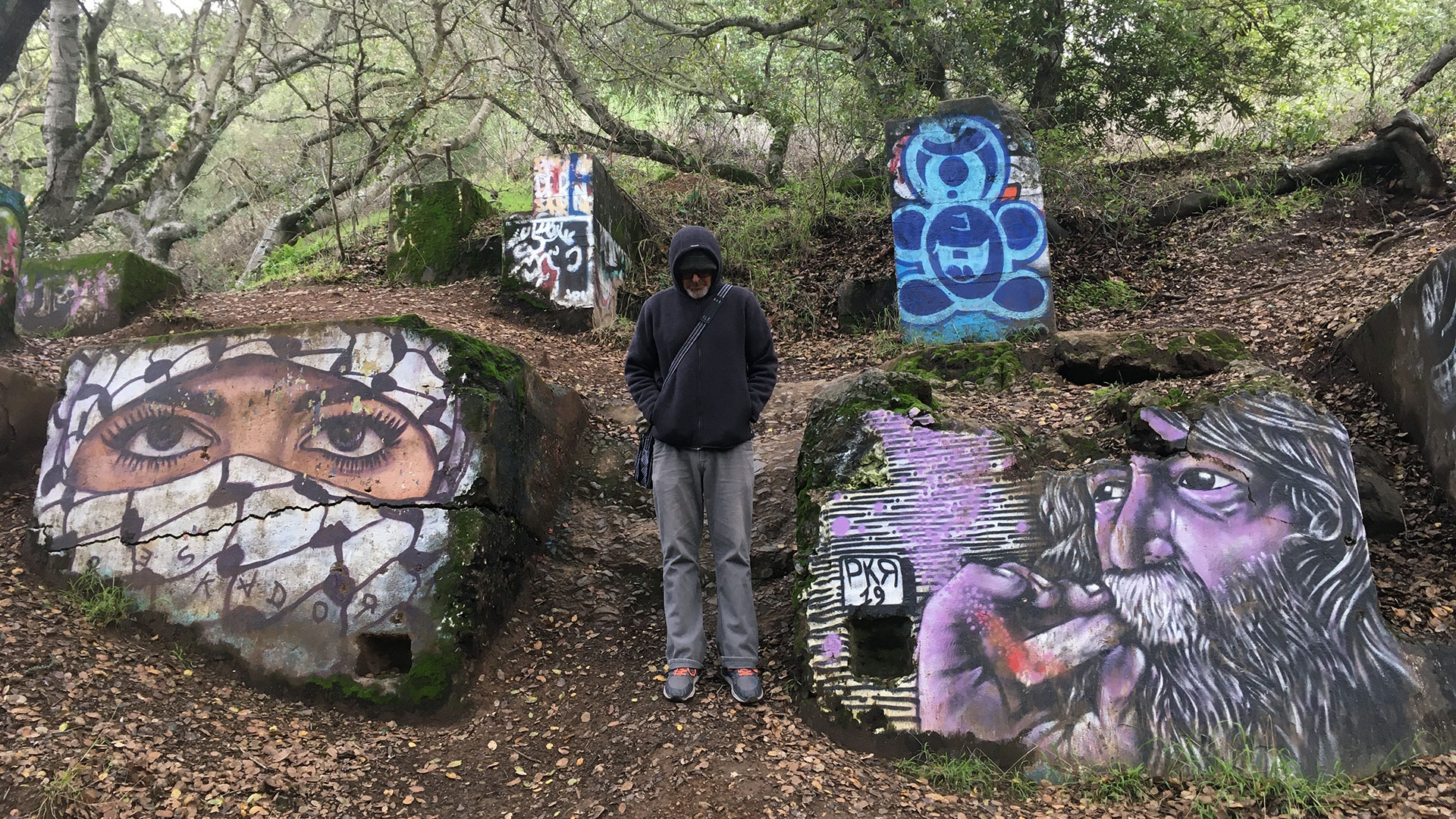 A man in black hoodie stands center, around him are remnants of concrete walls painted with vibrant art.