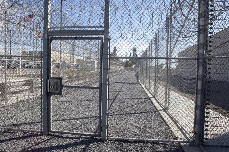 A chain-link fence with barbed wire at the top of a detention facility. An American flag waves in the distance.