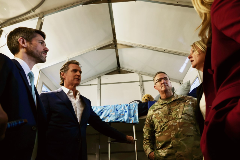 Two men in suits, including Gov. Newsom, consult with a group of people in a tent.