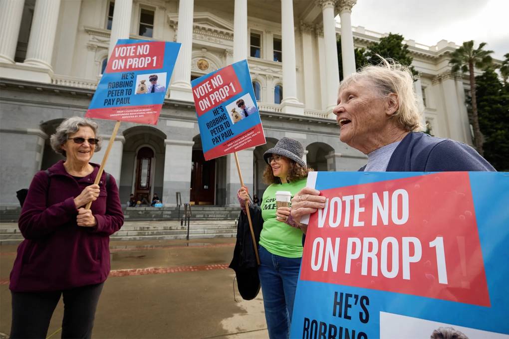 three woman stand outside a capitol building holding signs that say 'Vote No on Prop 1"