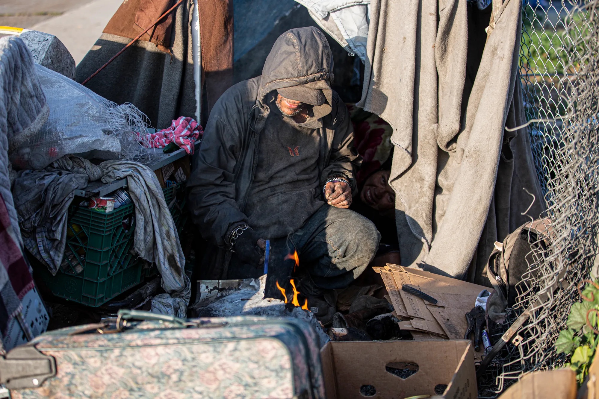 A seated man wearing a hooded jacket starts a fire in his makeshift tent.