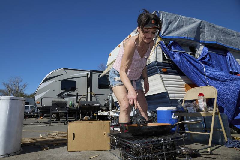A woman wearing a visor, light sleeveless top and jean shorts holds a utensil to cook food on a grill outside.