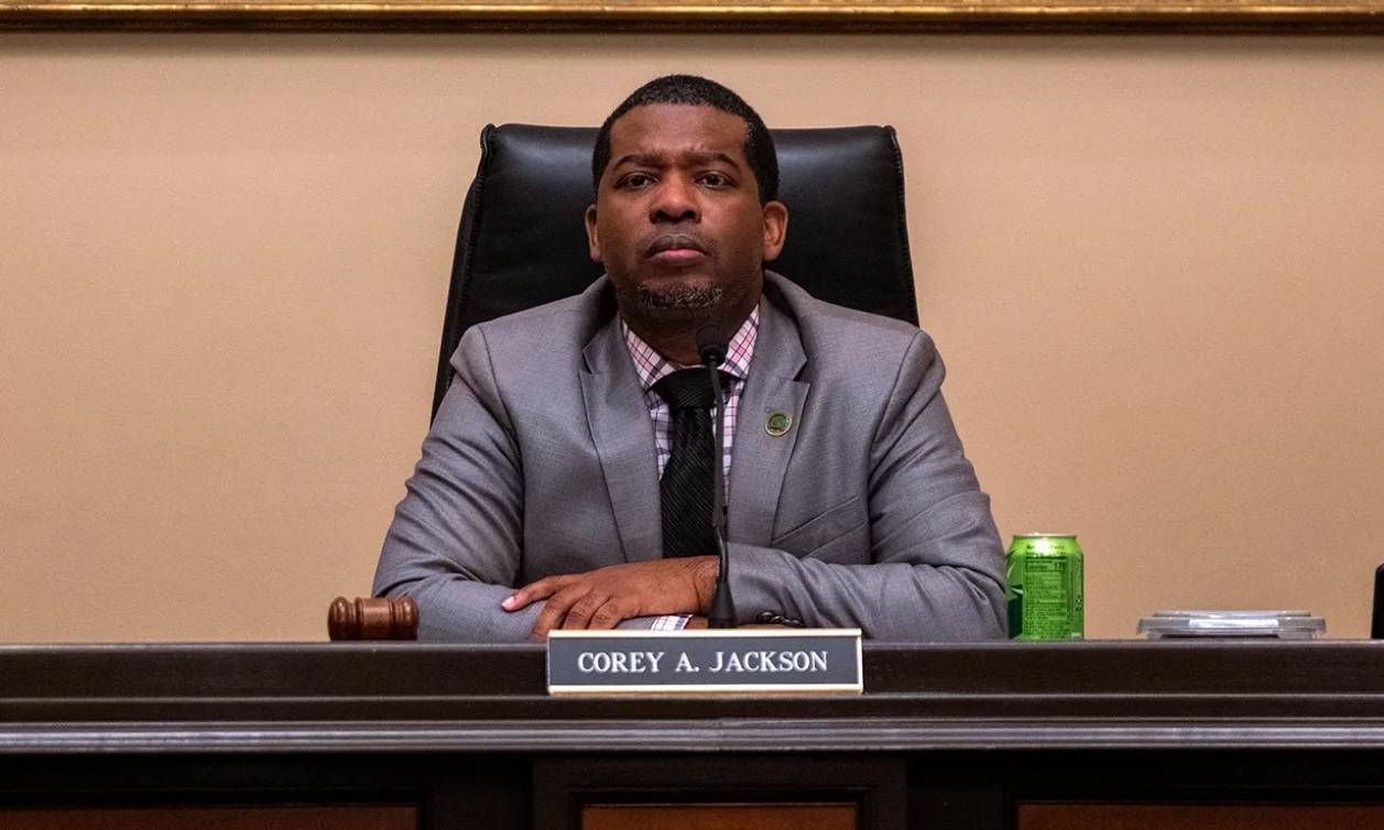 An African American man in a gray suit with black tie listens from behind a desk with his name on it.