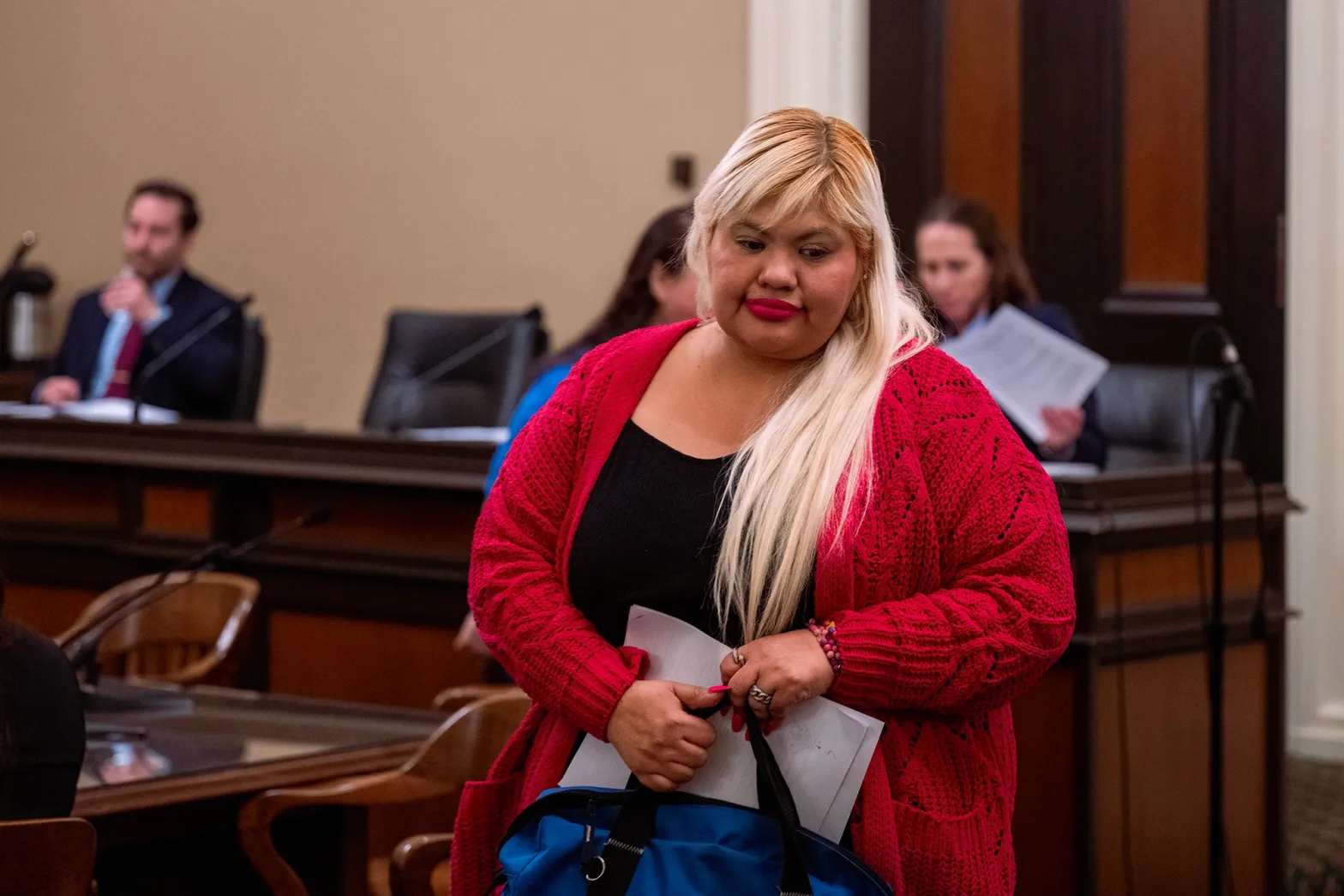 A woman with blond hair and a red cardigan stands in court, looking toward the floor.