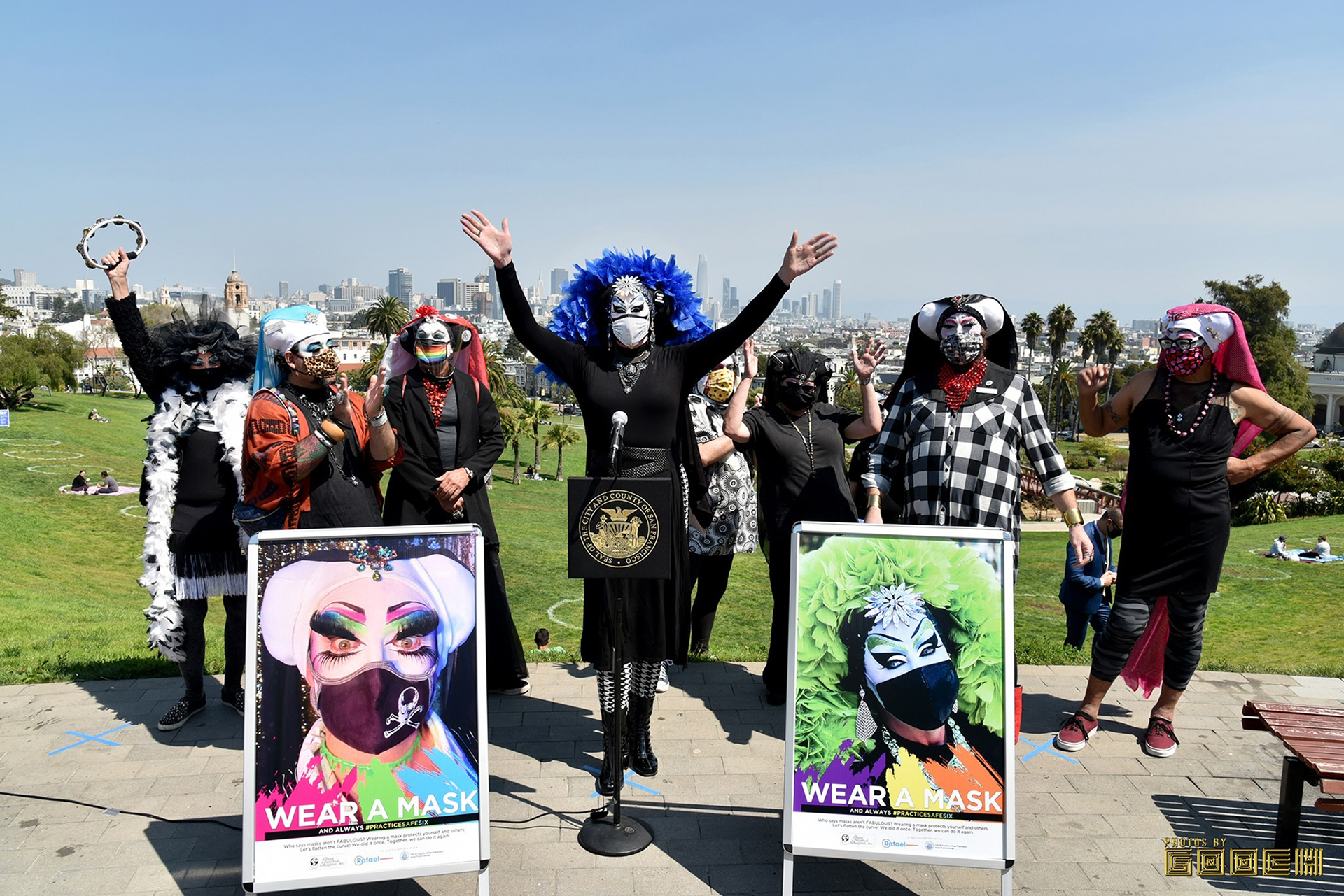 Seven "sisters" in their drag nun attire stand in front of Dolores Park in San Francisco. Near them is a sign that says "wear a mask." They are all wearing masks as well.