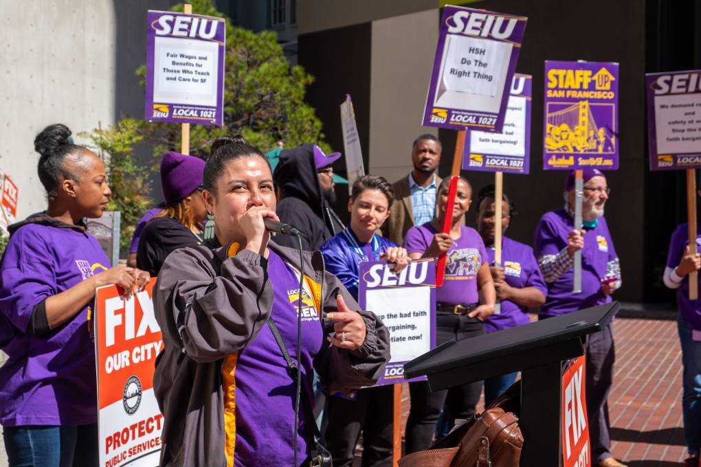 A group of people in purple shirts hold signs while standing in front of a building. One woman speaks into a microphone.