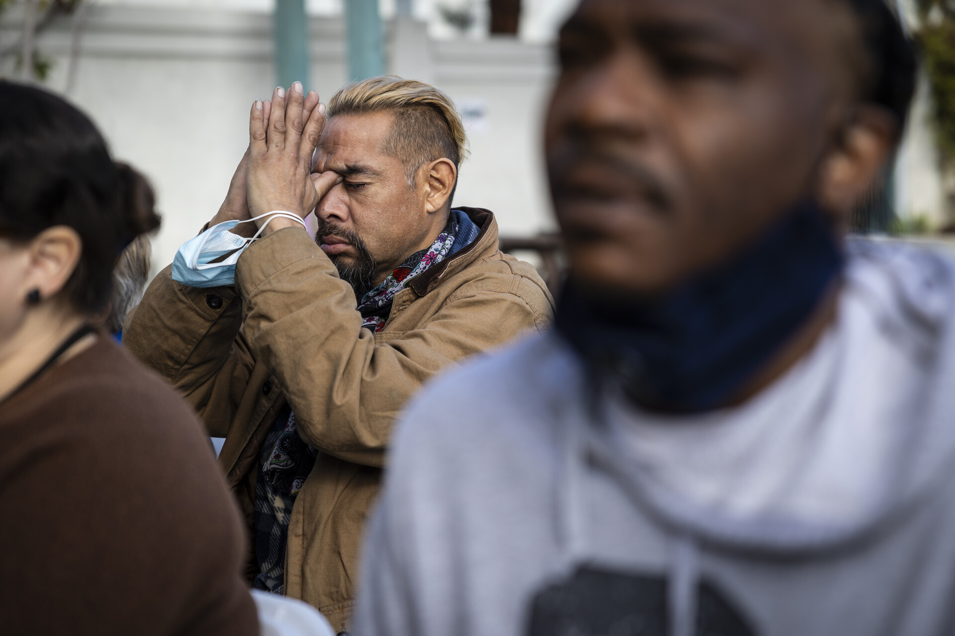 A man prays with his hands held up, behind others, outside.