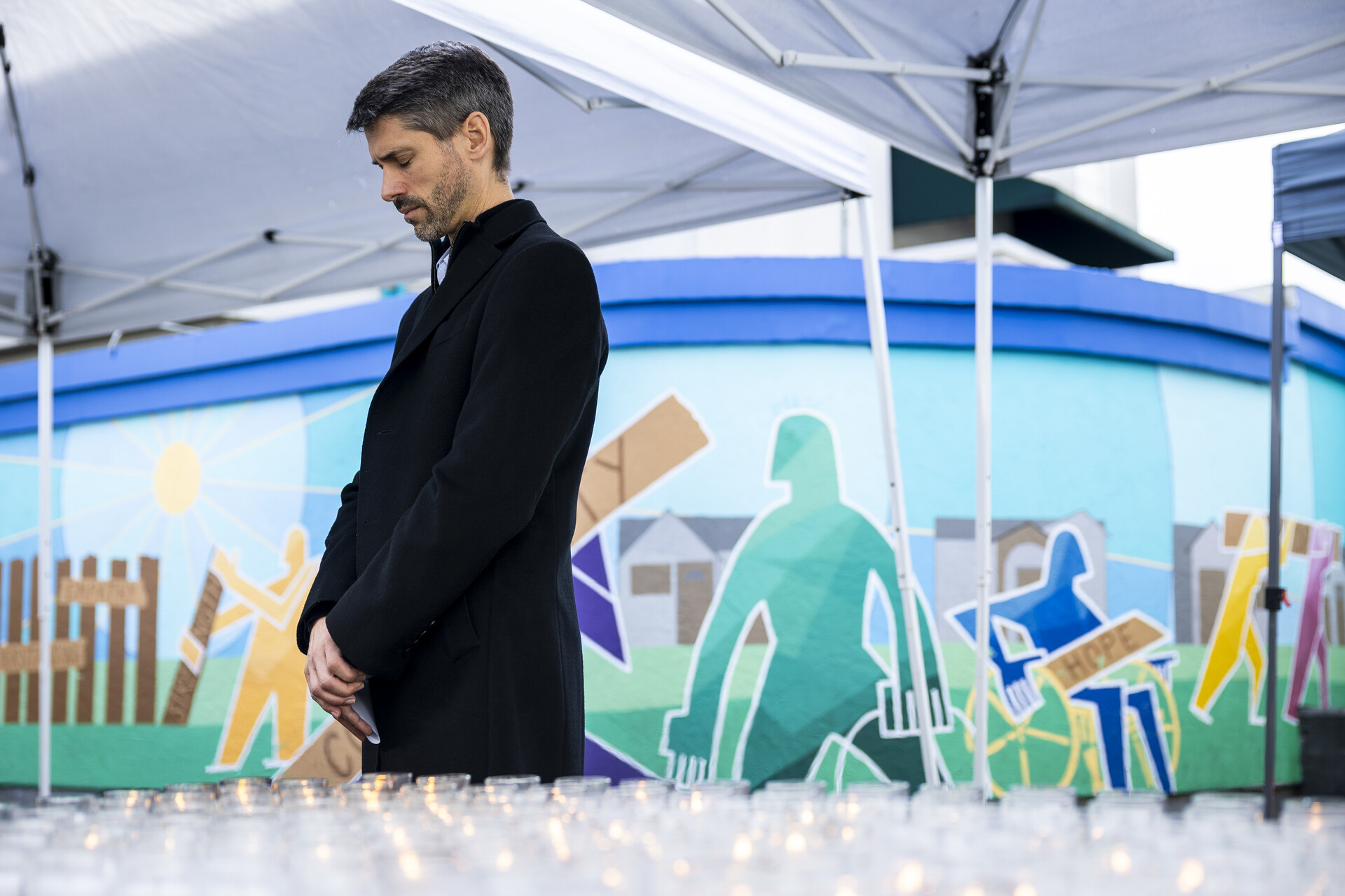A white middle-aged man stands in a moment of silence with mural behind him outdoors under a tent.