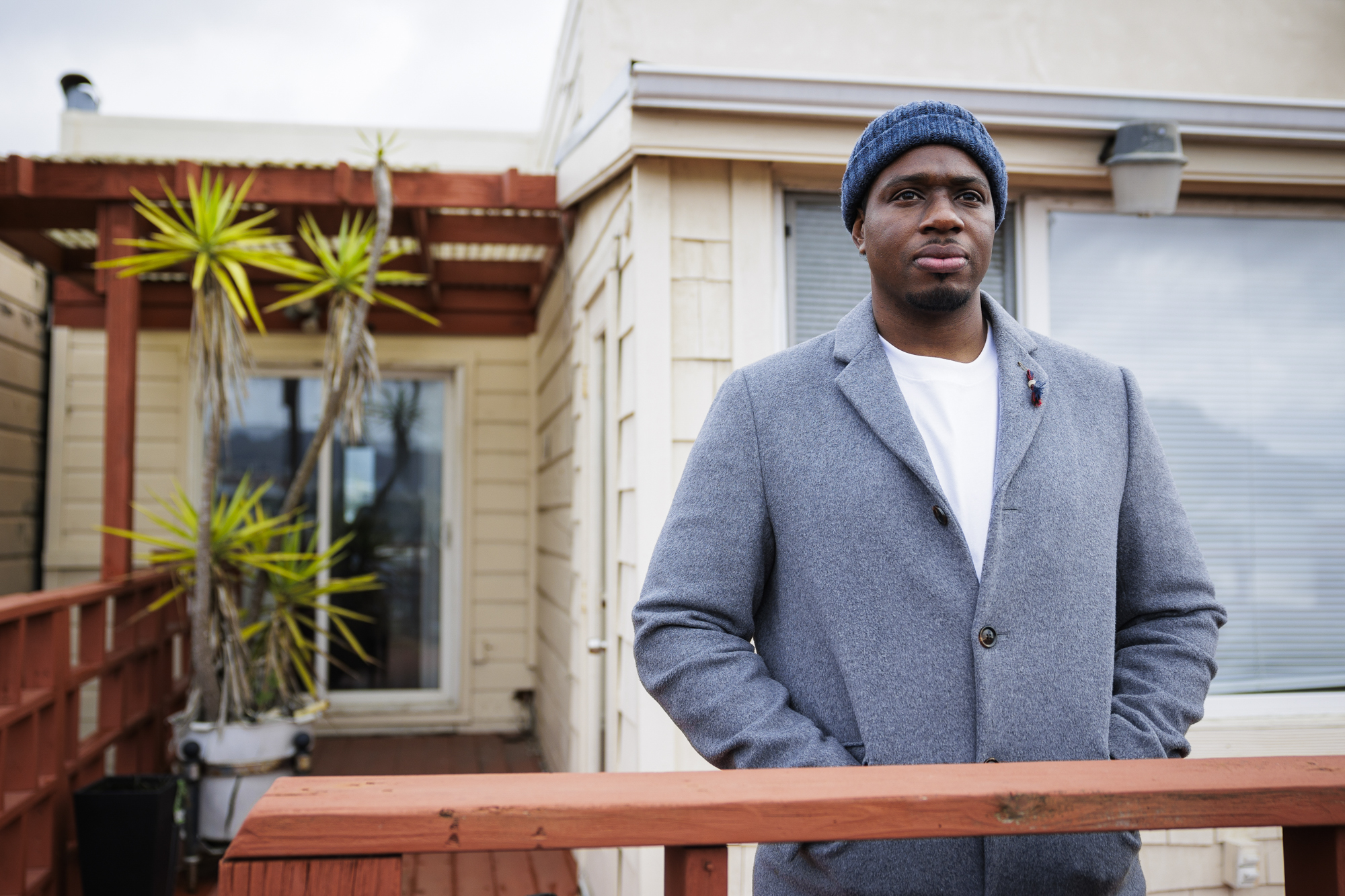 An African American man with a gray blazer and white shirt and a blue beanie stands in front of a house looking away from camera.