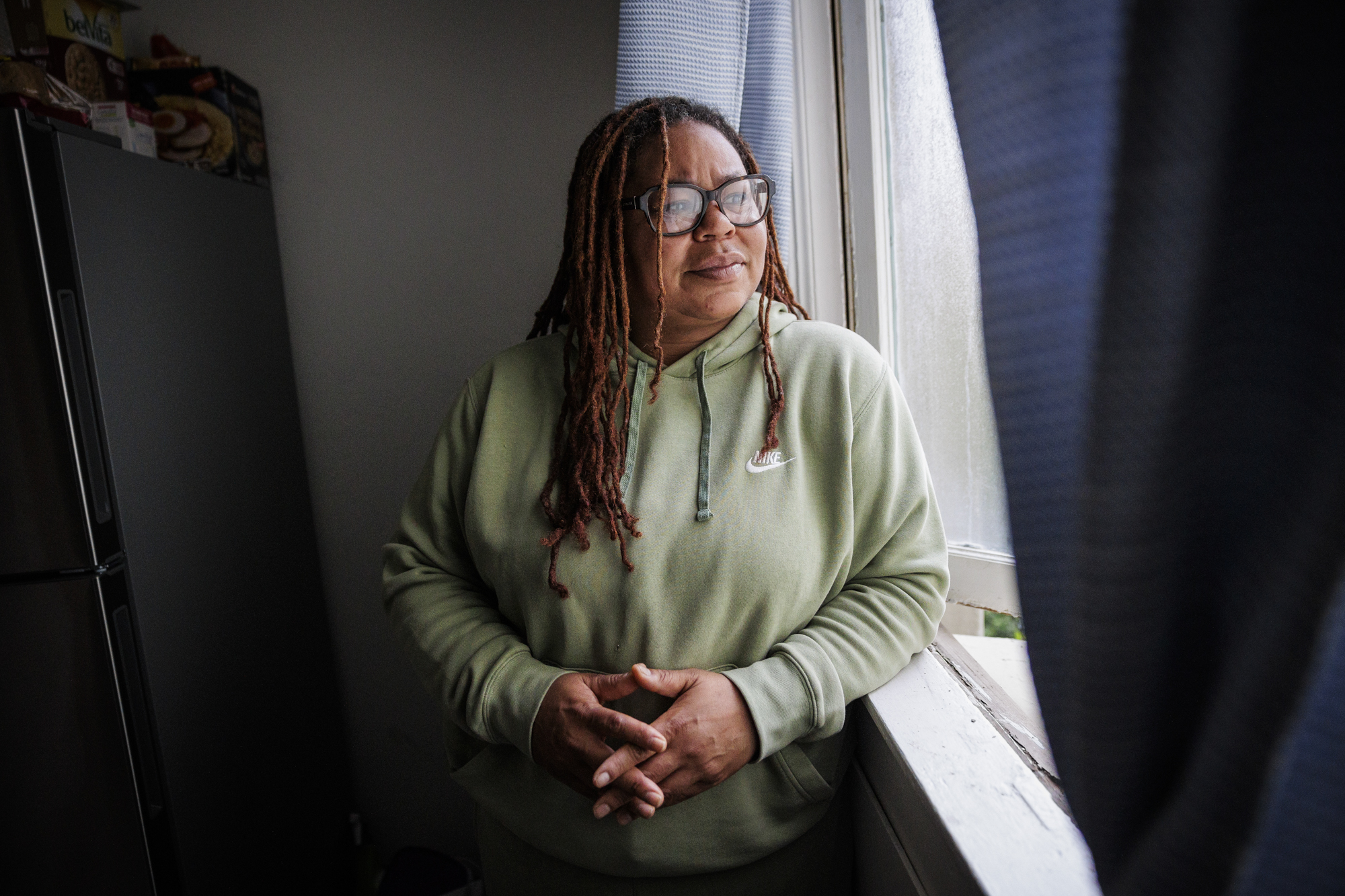 A middle-aged African American woman with long braided hair and glasses looks out from a window.