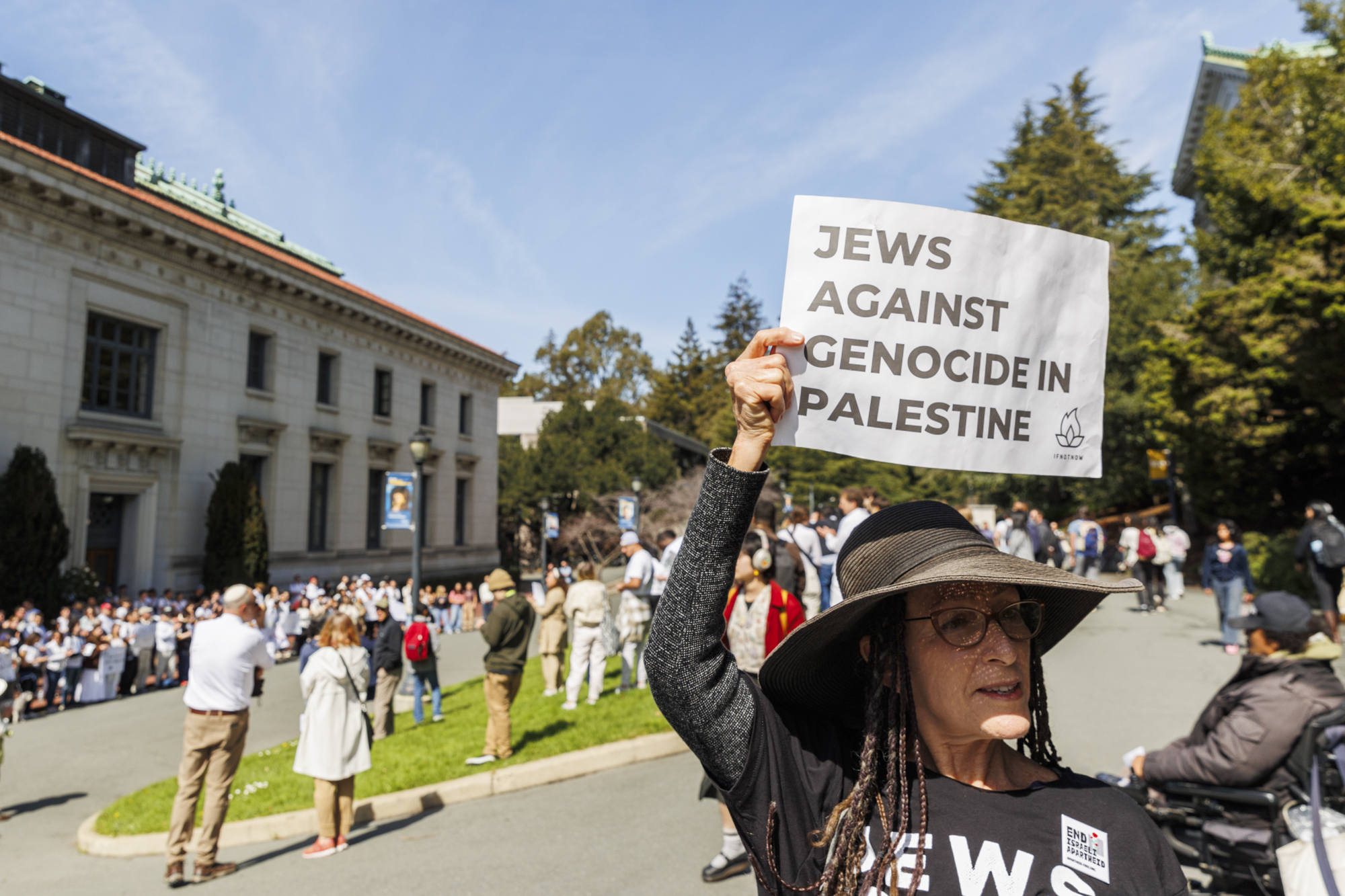 A person stands in an outdoor setting in front of a large group of people holding a sign that reads "Jews Against Genocide in Palestine".