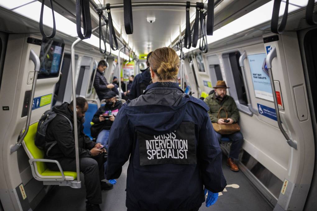 A woman in a dark-blue jacket that says 'Crisis Intervention Specialist' on the back walks through a BART train.