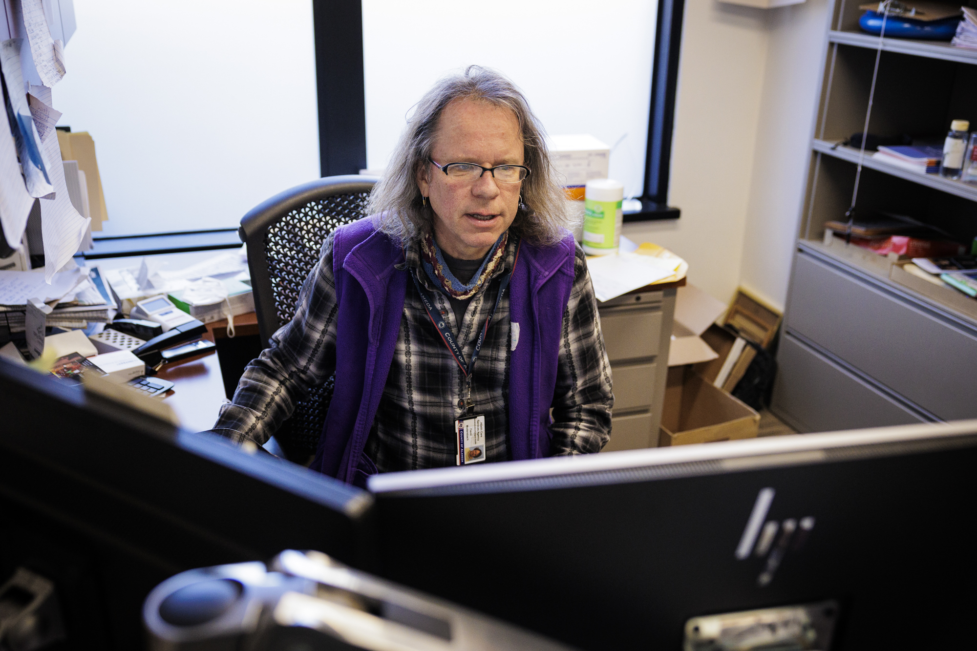 a man with glasses and long hair, wearing a flannel shirt, sits behind a computer