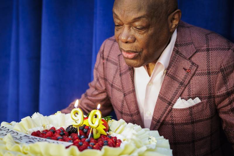 An older African American man in a suit and white shirt bends over to blow out candles on a birthday cake with the figure 90 on it.