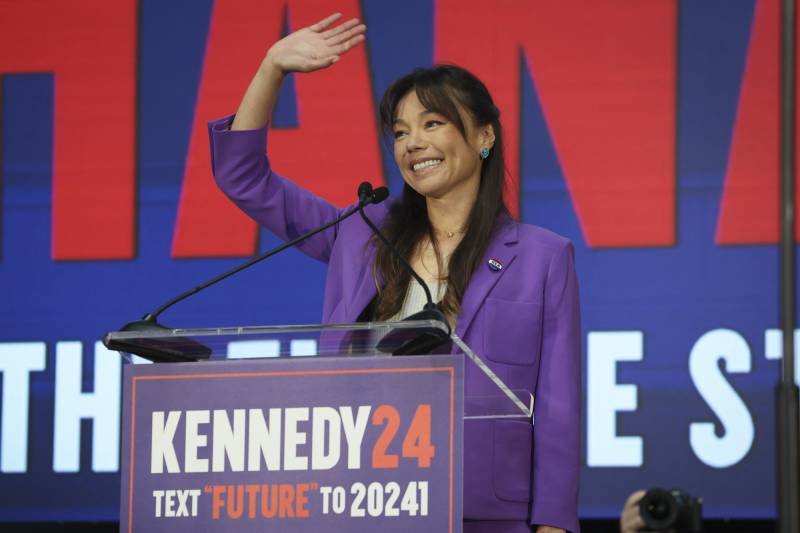 An Asian American woman with long hair and a purple suit with a white shirt waves from a stage behind a lectern that reads "Kennedy 24."