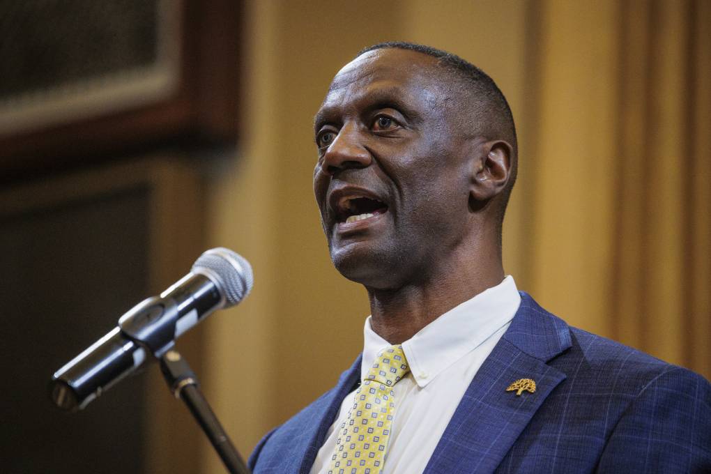 An African American man with a blue suit and yellow patterned tie speaks into a microphone.