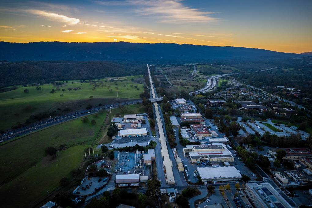An aerial view of a very long, skinny and beige building that extends toward the horizon line. It is sunset, and in the background are the Santa Cruz mountains.
