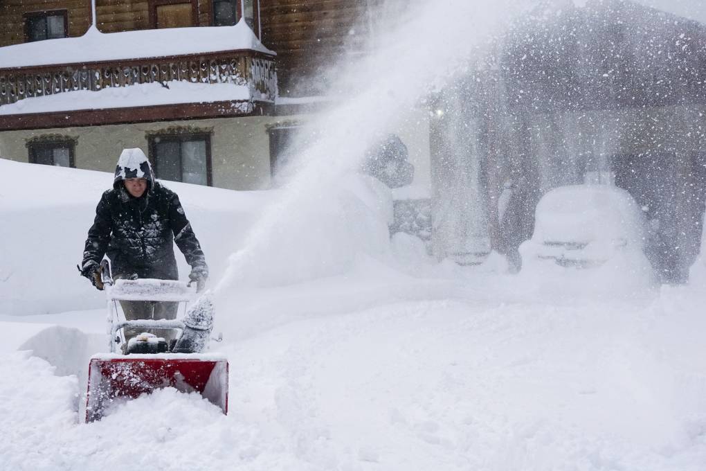 A man wearing a snow jacket uses a snow blower to clear snow outside of a house.