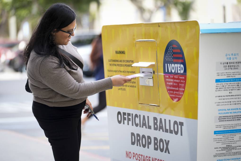 A woman with dark brown hair drops a ballot into a ballot box.