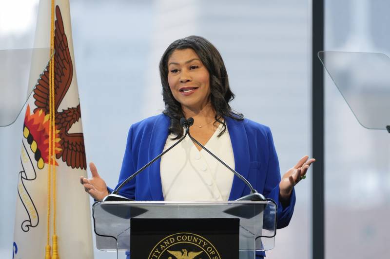 Mayor London Breed, wearing a blue jacket, delivers a speech in front of a podium, with teleprompters or either side of her.