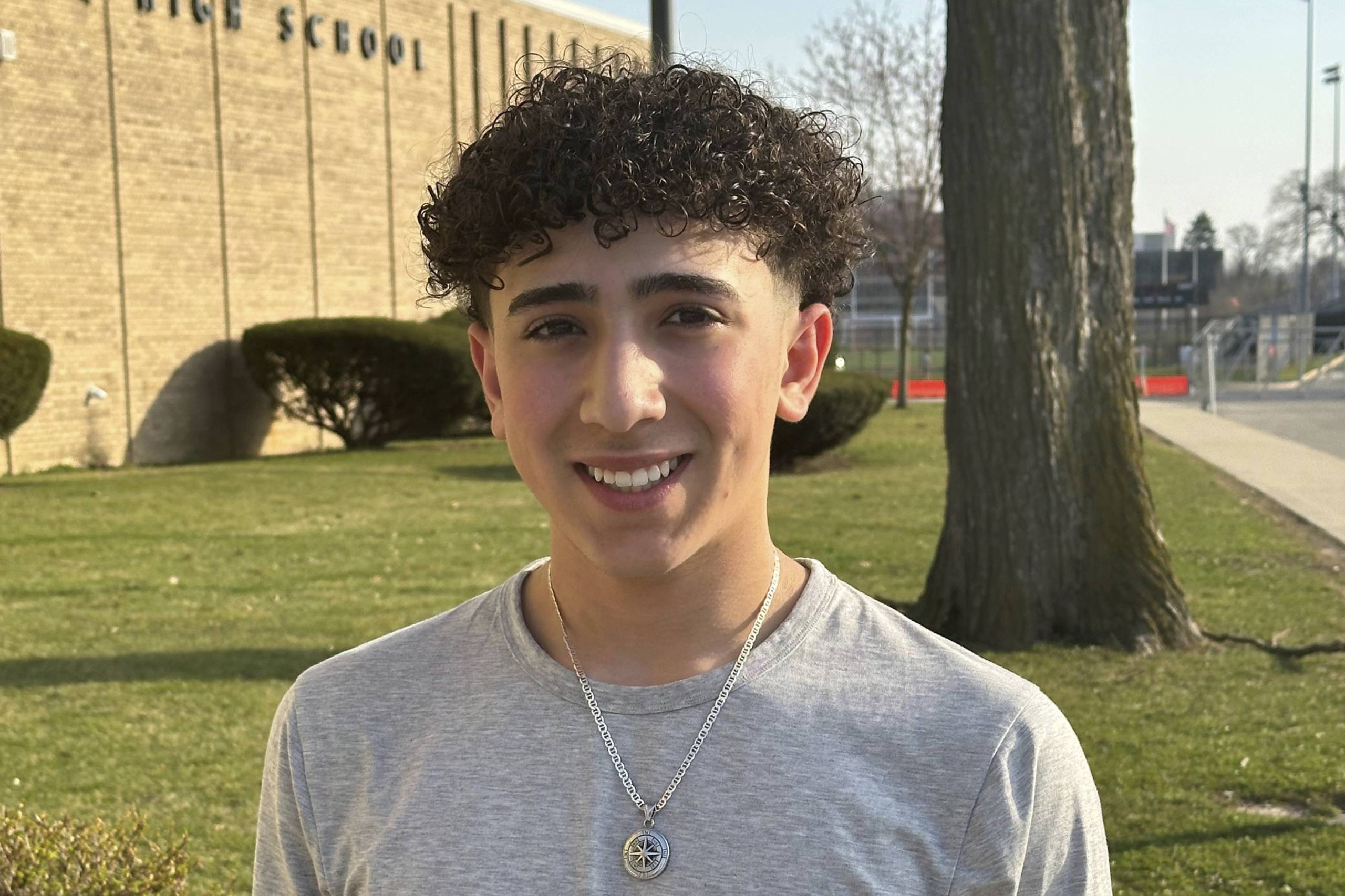 A 9th grade student, male, wearing gray shirt, smiles at the camera with a school building and lawn with trees behind him.