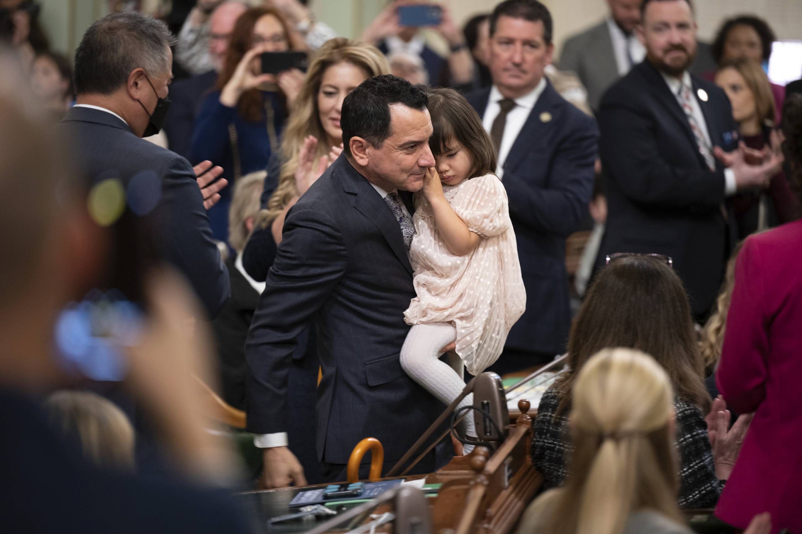 A man in a blue suit holds his young daughter in his arms as he passes lawmakers on his way to a lectern.
