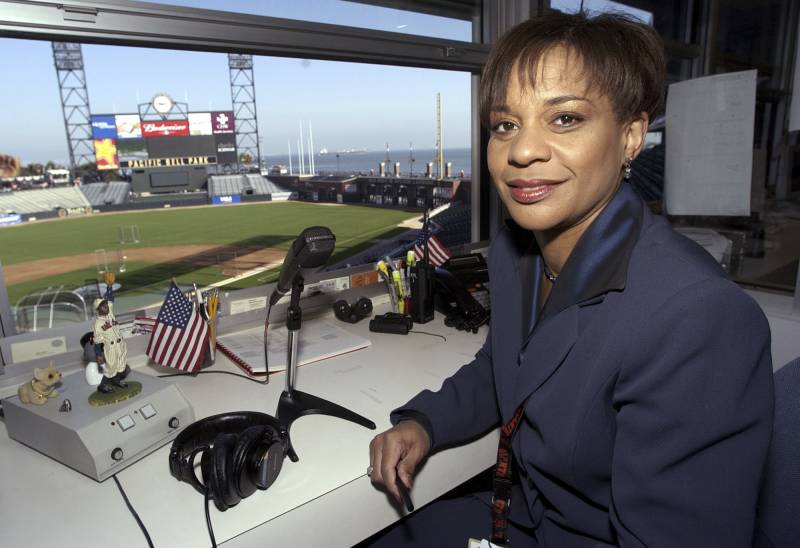 A black woman sits in a booth at a baseball stadium, in front of a microphone