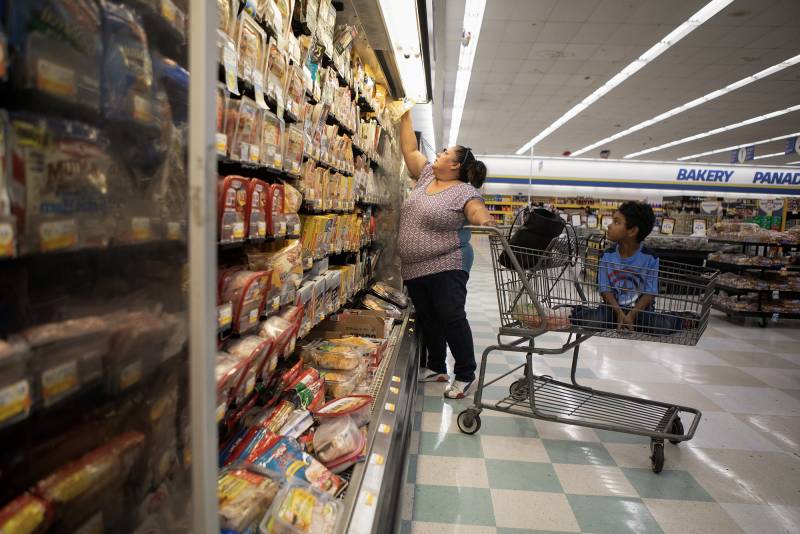 A woman reaches for an item on the top shelf at a grocery store beside a shopping cart with a child sitting in it.
