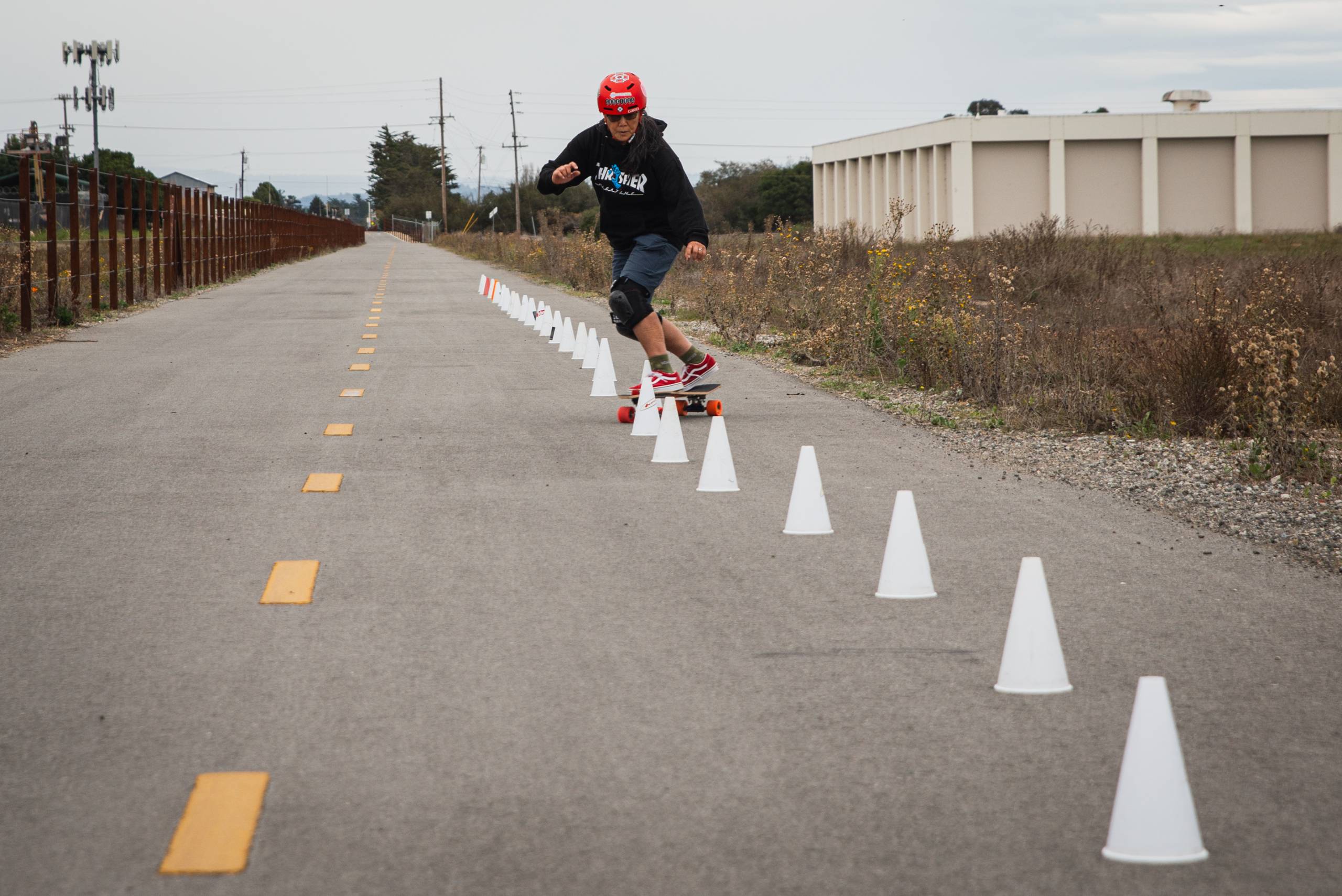A skateboarder with a red helmet slaloms through a line of white cones on a road.
