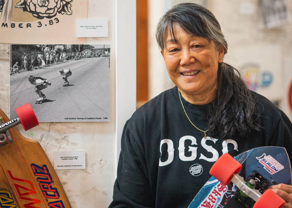 An older Asian woman is holding a skateboard and smiling, with old photos behind her.