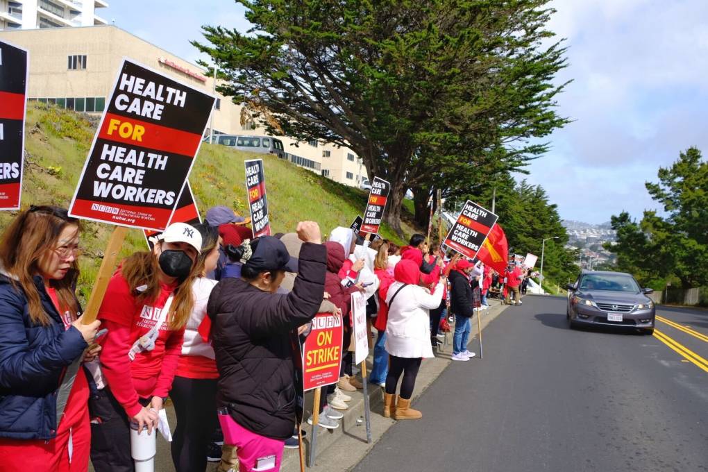 People stand on the side of the road holding black and red signs that say 'Health care for health care workers.'