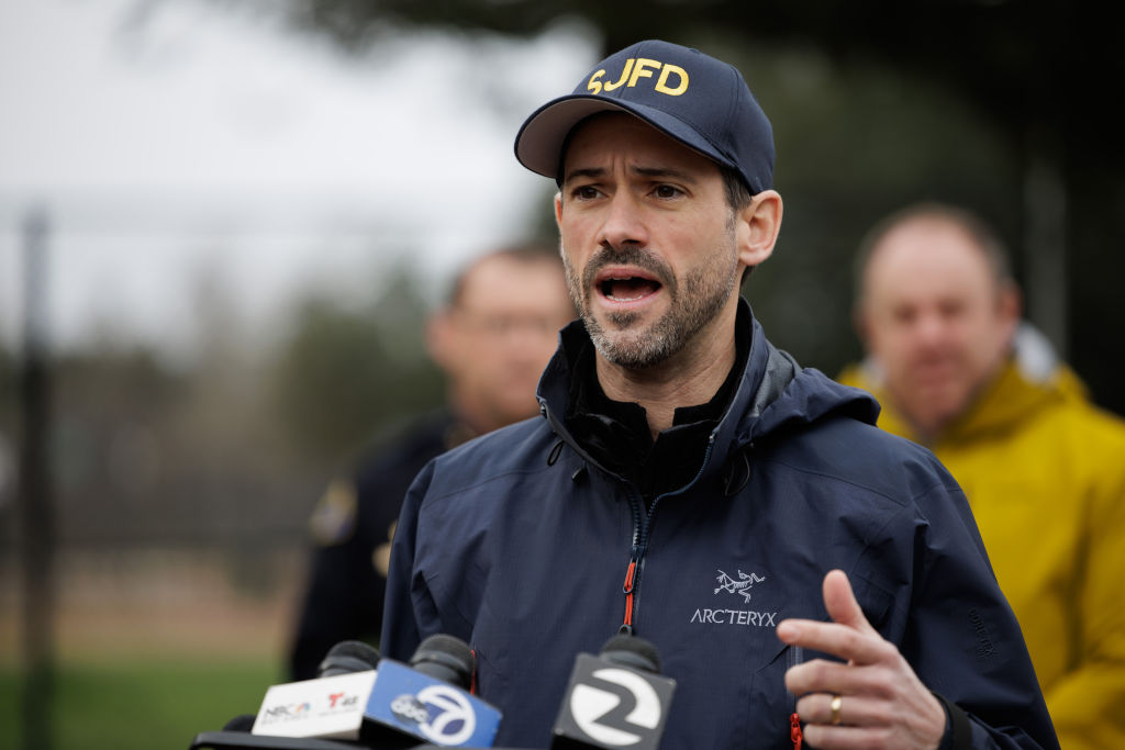 A middle-aged white man with a hat that reads "SJFD" speaks at a press conference outdoors, with two blurred men behind him.