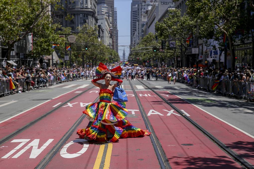 A drag queen stands in the middle of Market Street in San Francisco wearing a flamboyant gown with vibrant rainbow colors.